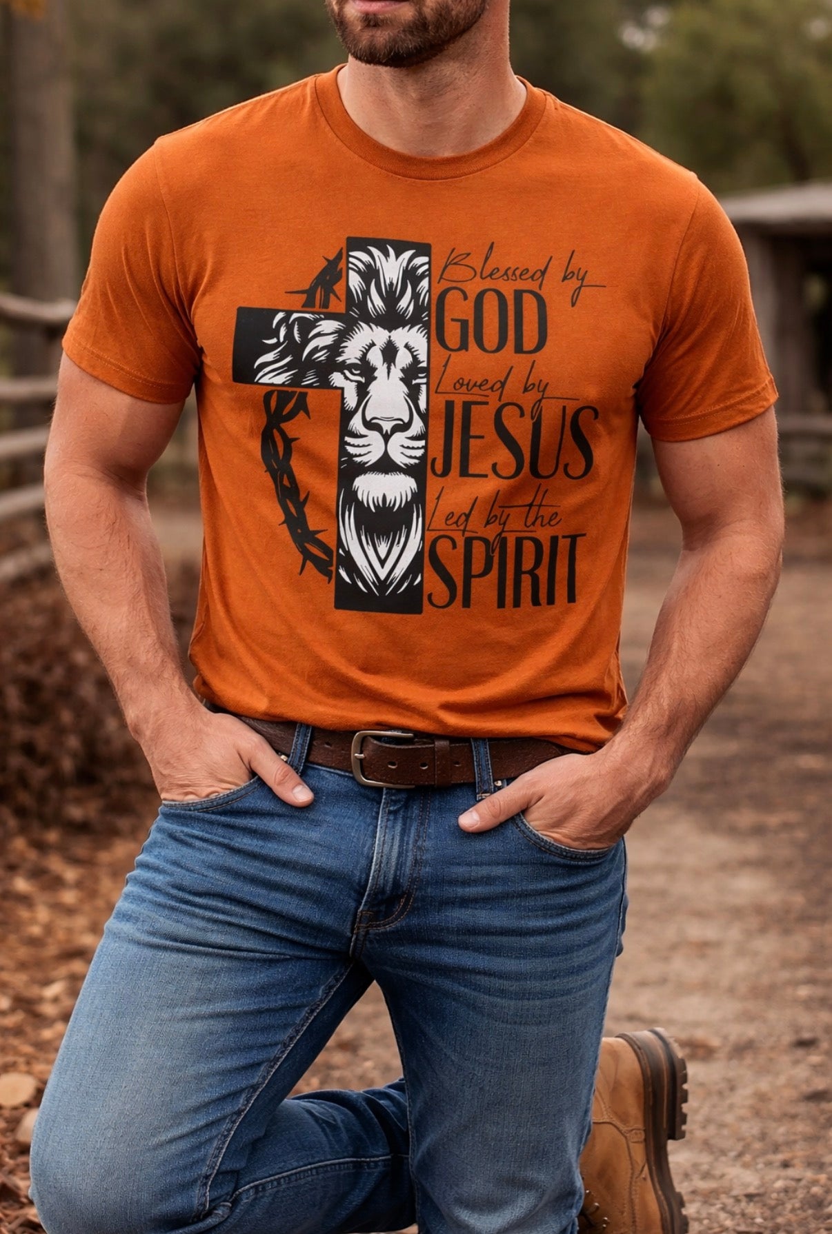 Man wearing a Texas Orange Christian T-Shirt with a lion and cross design and the phrase Blessed by God, Loved by Jesus, Led by the Spirit, paired with jeans and boots outdoors.